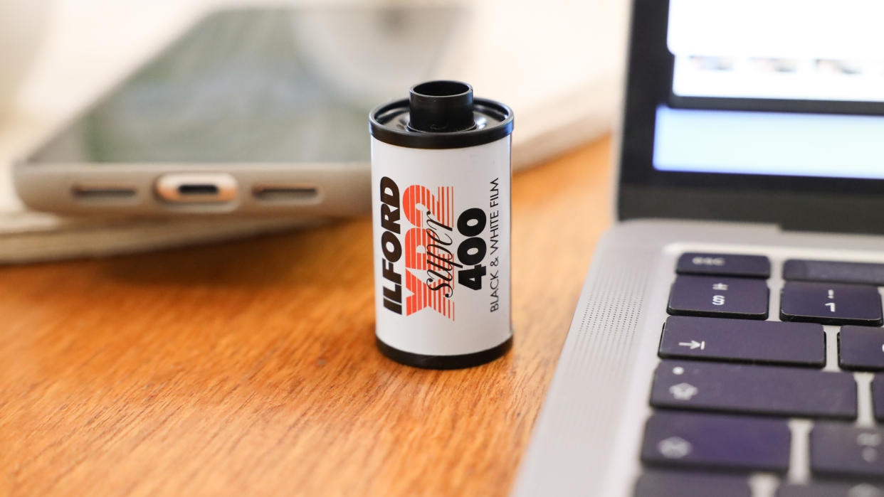  Ilford XP2 Super 35mm film canister on a wooden surface next to a MacBook. 