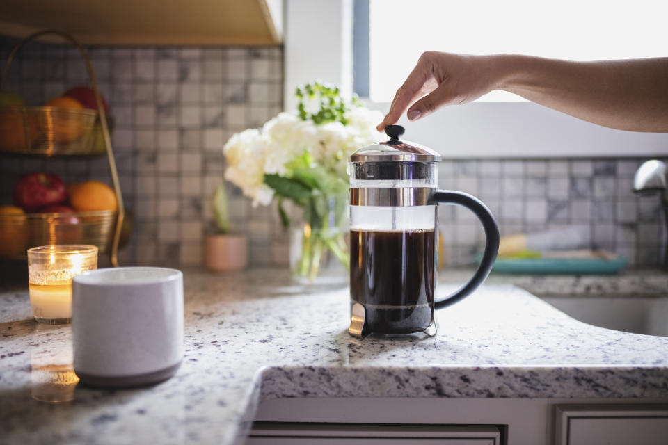 Hand pressing down the plunger of a French press coffee maker on a kitchen countertop with a lit candle, fruit basket, and flowers in the background