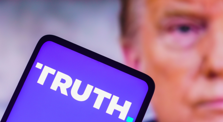 In this photo illustration, the Truth Social logo seen displayed on a smartphone with a photo of former US President Donald Trump displayed in the background. DJT stock