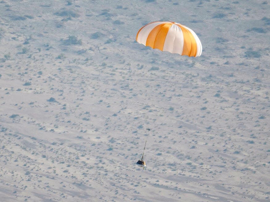 A black container attached to an orange-and-white parachute over the Utah desert