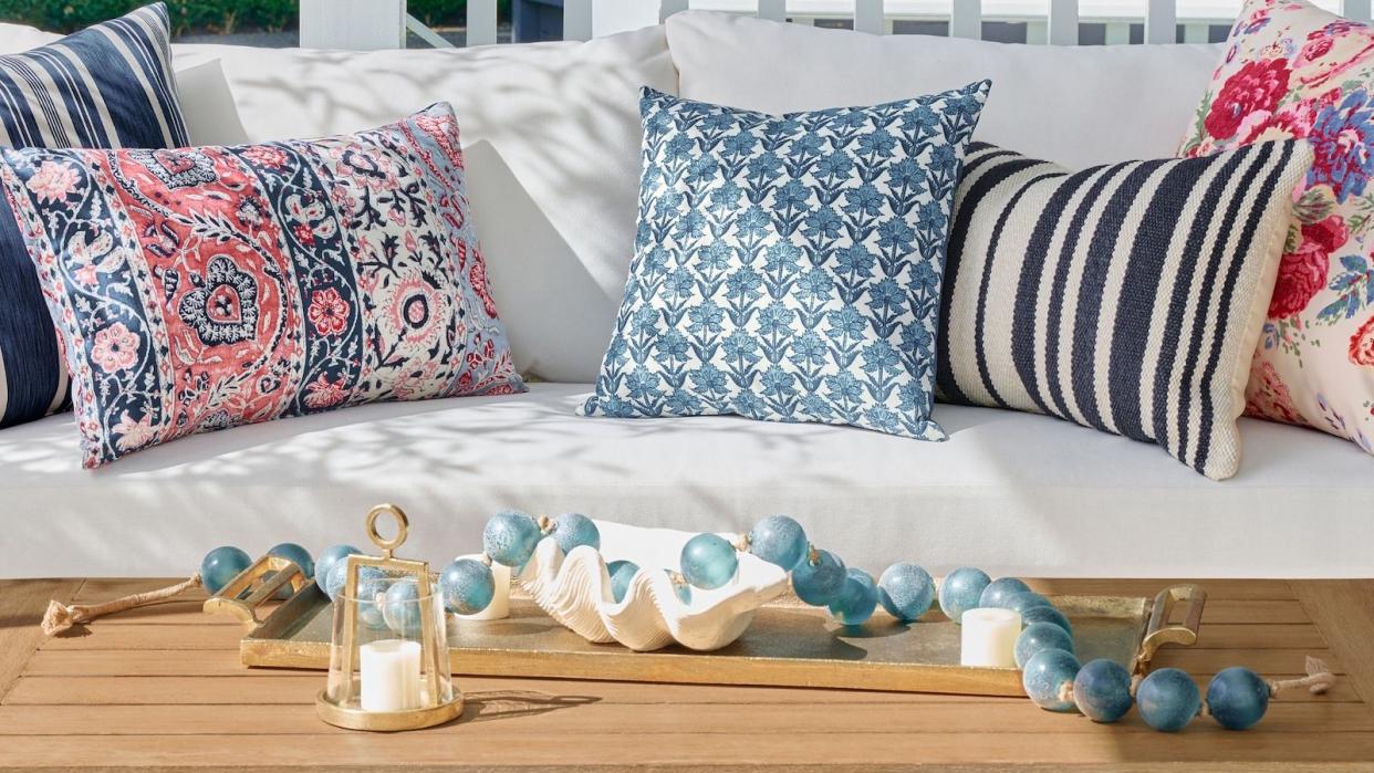  Pottery Barn outdoor white cushioned couch with multicolored blue and pink outdoor pillows next to a wooden table with coastal decor . 