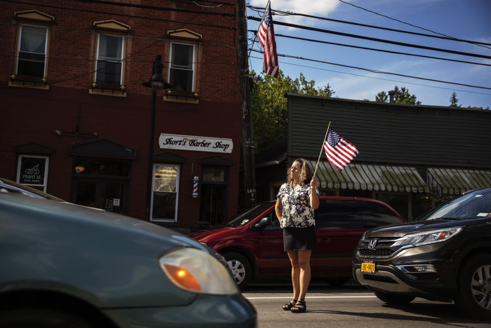 Sarah Short stands outside her barber shop to watch a memorial procession pass through Trumansburg, N.Y., Saturday, Aug. 31, 2019, for Sgt. James Johnston, who was killed in Afghanistan in June. On this late summer Saturday, a procession of fire engines, motorcycles and squad cars were greeted by clusters of flag-waving folks. With its celebration of Johnston, the war came home to this hamlet in upstate New York. (AP Photo/David Goldman)