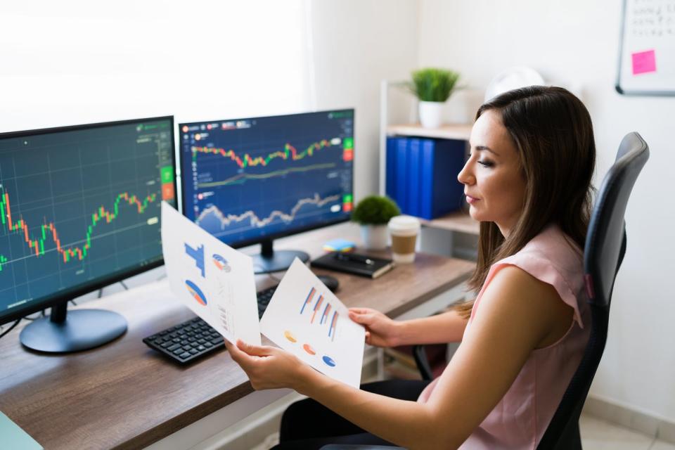 A person reviews charts on paper while sitting in front of computer screens with more stock charts.