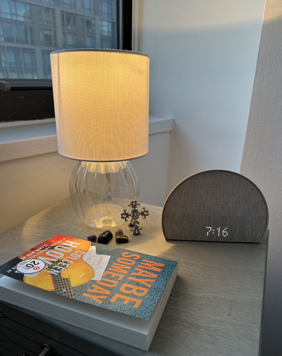 Table with lamp, alarm clock reading 9:16, and a book titled "Maybe Someday". Decor includes earrings and stones