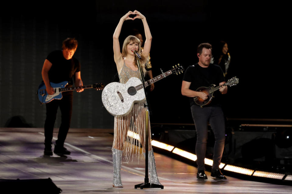 Taylor Swift performing with a fringed dress and guitar, alongside band members on stage doing heart hands