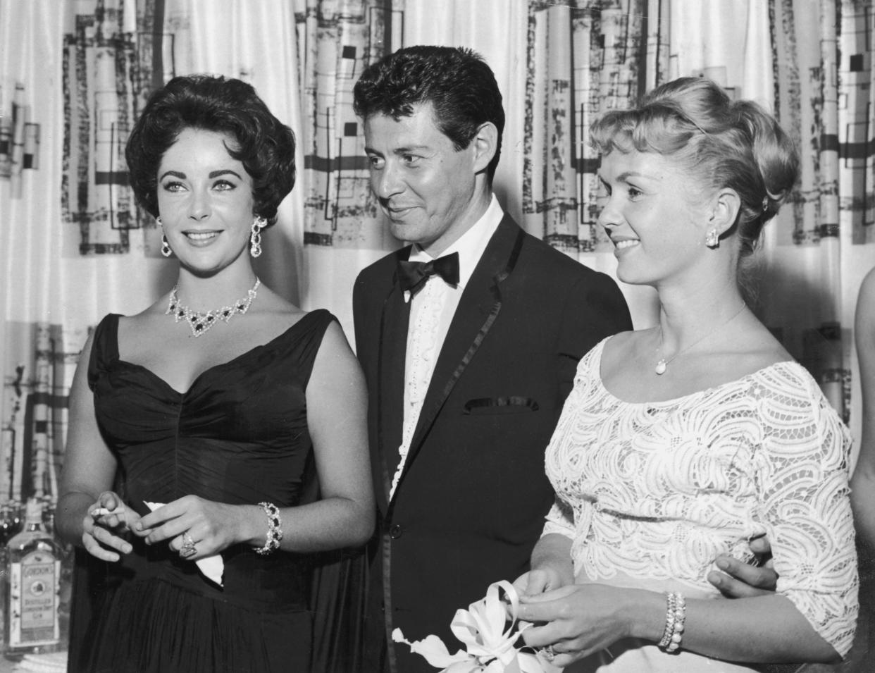 1958: el cantante estadounidense Eddie Fisher, vestido con un esmoquin, se para con el brazo alrededor de su esposa, la actriz estadounidense Debbie Reynolds (derecha) y sonríe mientras mira a la actriz británica Elizabeth Taylor, fumando un cigarrillo, Las Vegas, Nevada. Al año siguiente, Fisher dejó Reynolds y se casó con Taylor. (Foto de Hulton Archive/Getty Images)