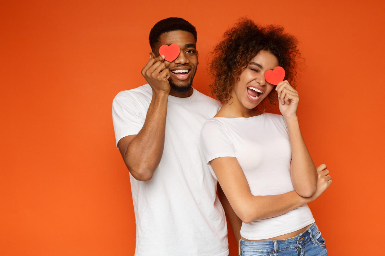 Love and happiness concept. Cheerful enjoyable young couple with little small hearts covering one eye, orange background