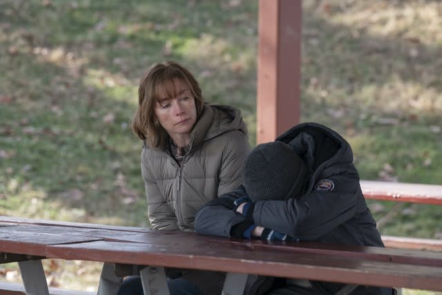 A woman sits at a picnic table looking on as a boy buries his head in his arms