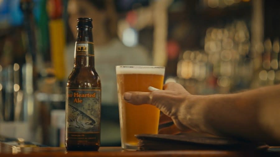 A man reaches for a glass of beer next to a bottle of Bell's Two Hearted Ale, showing its iconic trout logo.