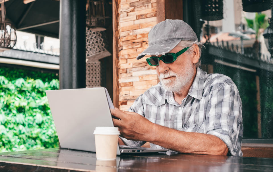 An older man with a white beard, wearing a plaid shirt, cap, and sunglasses, uses a laptop at an outdoor café table with a cup of coffee