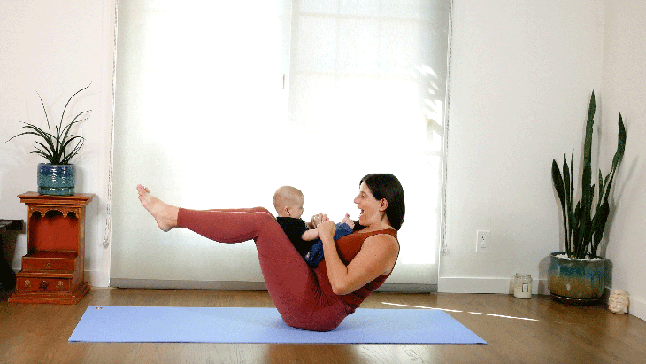 Woman sitting on your yoga mat leaning slightly back with her baby resting on her lap doing Boat Pose in yoga