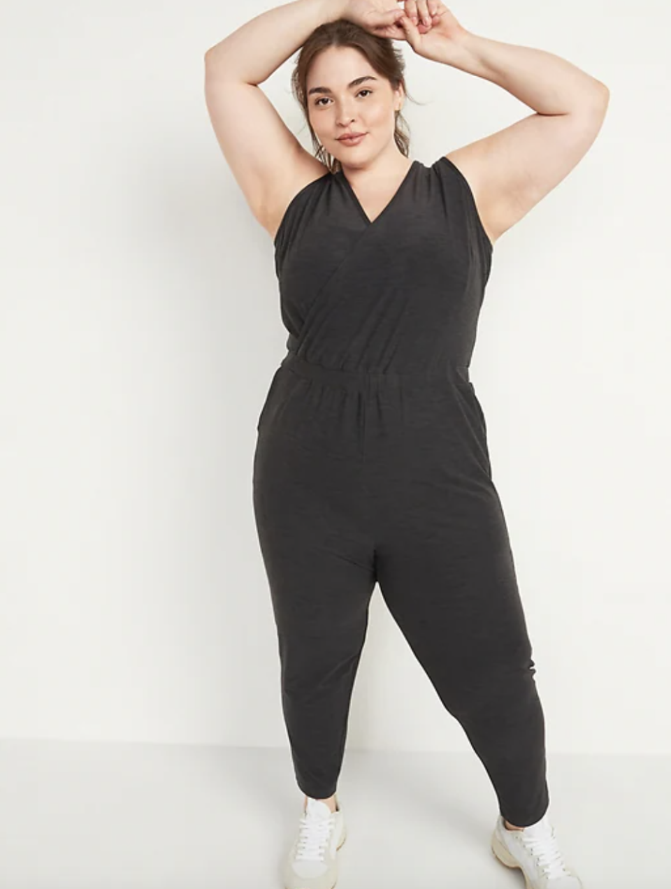 brunette plus size model posing in black sleeveless jumpsuit and white sneakers