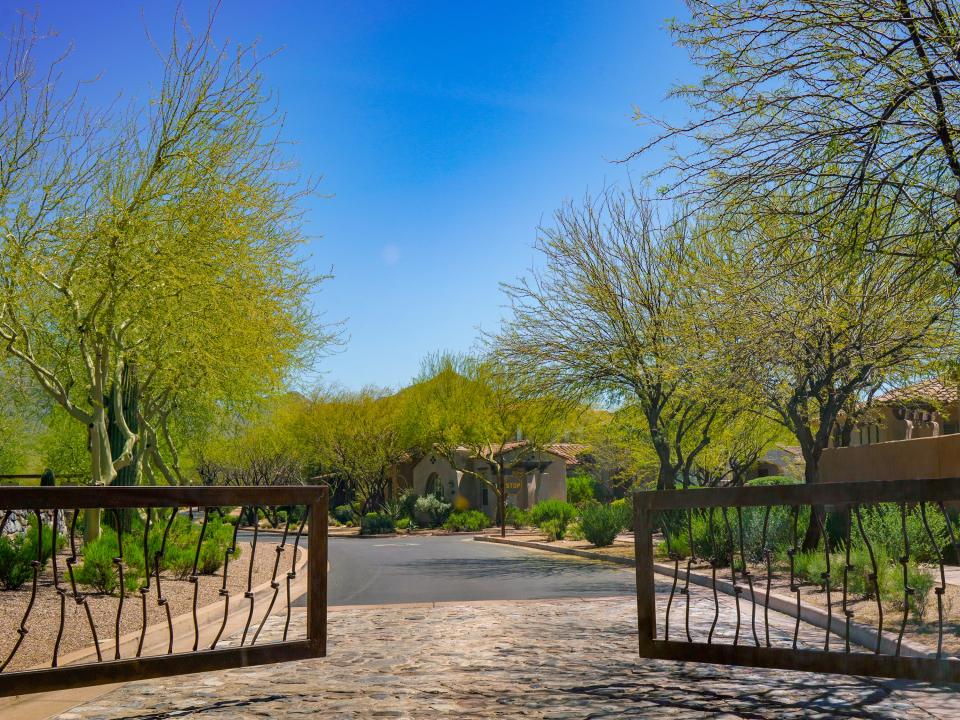Gates open into a neighborhood with adobe houses shaded by bushes and thin trees