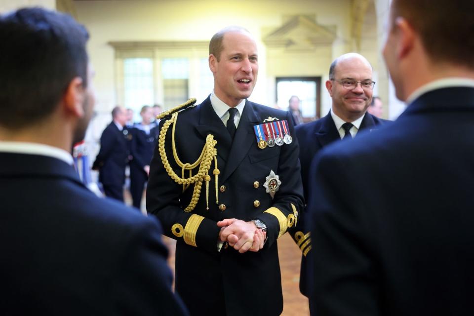 prince william stands in a naval uniform with several medals, he smiles and clasps his hands in front of him, a man stands to the right and two men face him