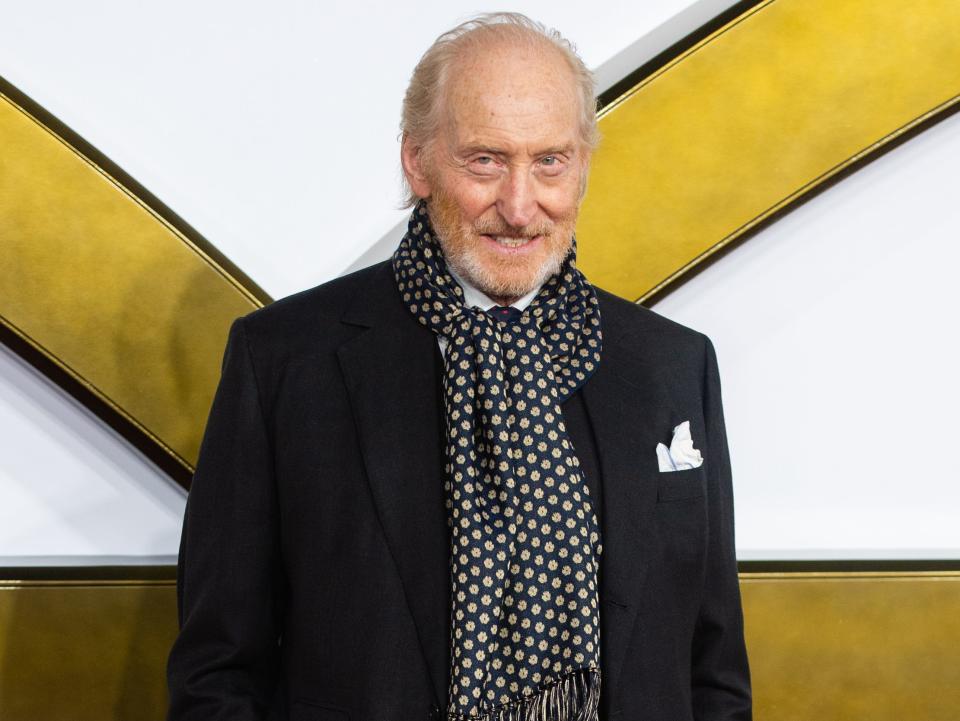 charles dance on the red carpet for the king's man, wearing a black suit coat, black and gold fringed scarf, and white pocket chief