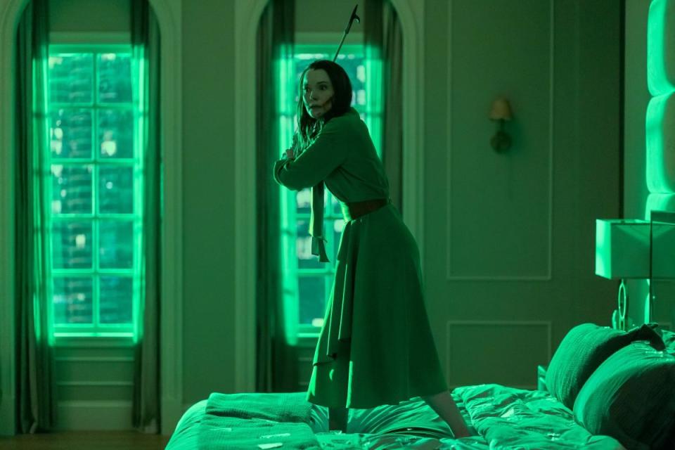 A woman holds a bat standing on a bed bathed in green light in The Fall Of The House Of Usher