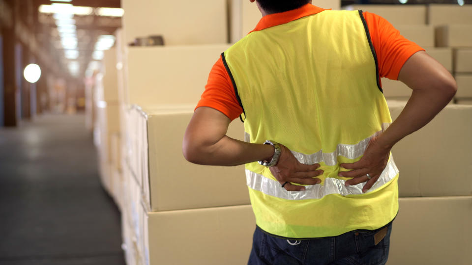 A warehouse worker wearing a high-visibility vest and an orange shirt holds their lower back, standing in front of stacked cardboard boxes