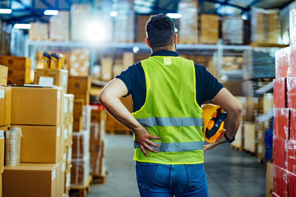 A worker wearing a high-visibility vest and holding a hard hat stands in a warehouse filled with stacked cardboard boxes. The worker is facing away