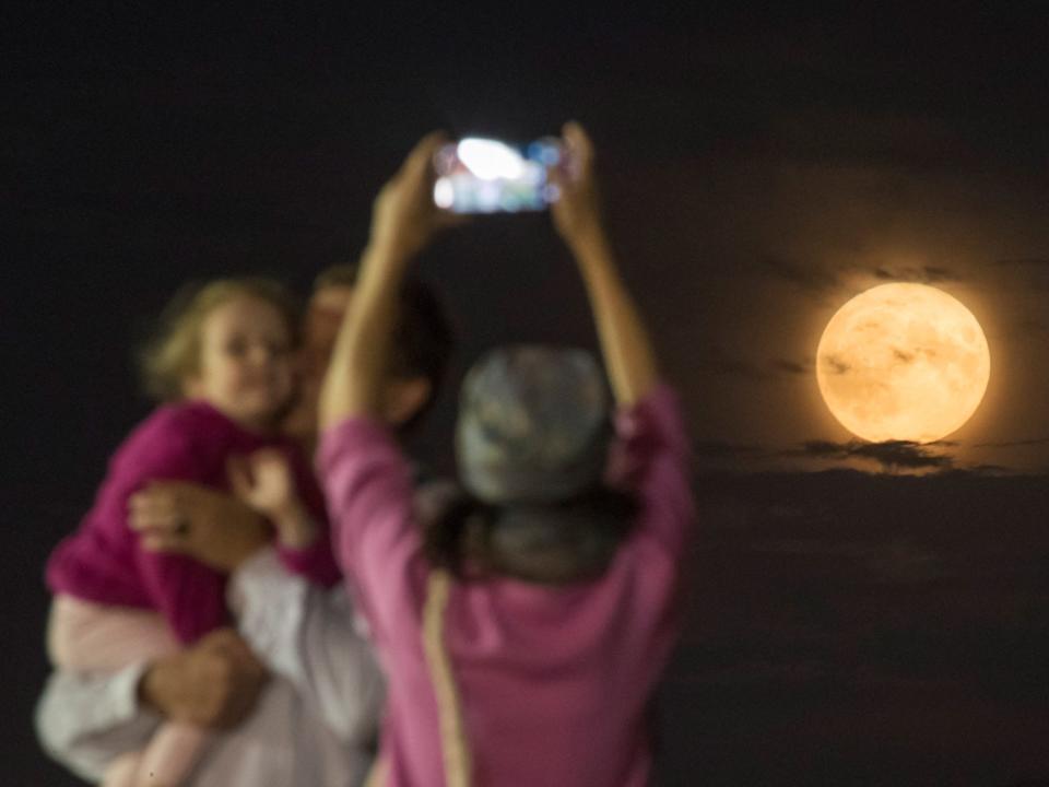 person takes phone photo of another person holding a child in front of yellow full moon