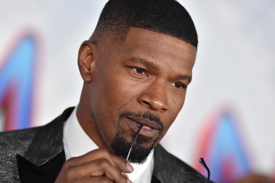 Jamie Foxx poses while biting onto a pair of glasses on the red carpet premiere of a Spider-Man film.