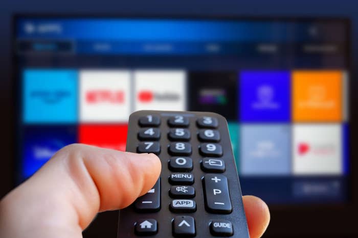 A hand holds a TV remote, pointing at a screen displaying various streaming service icons