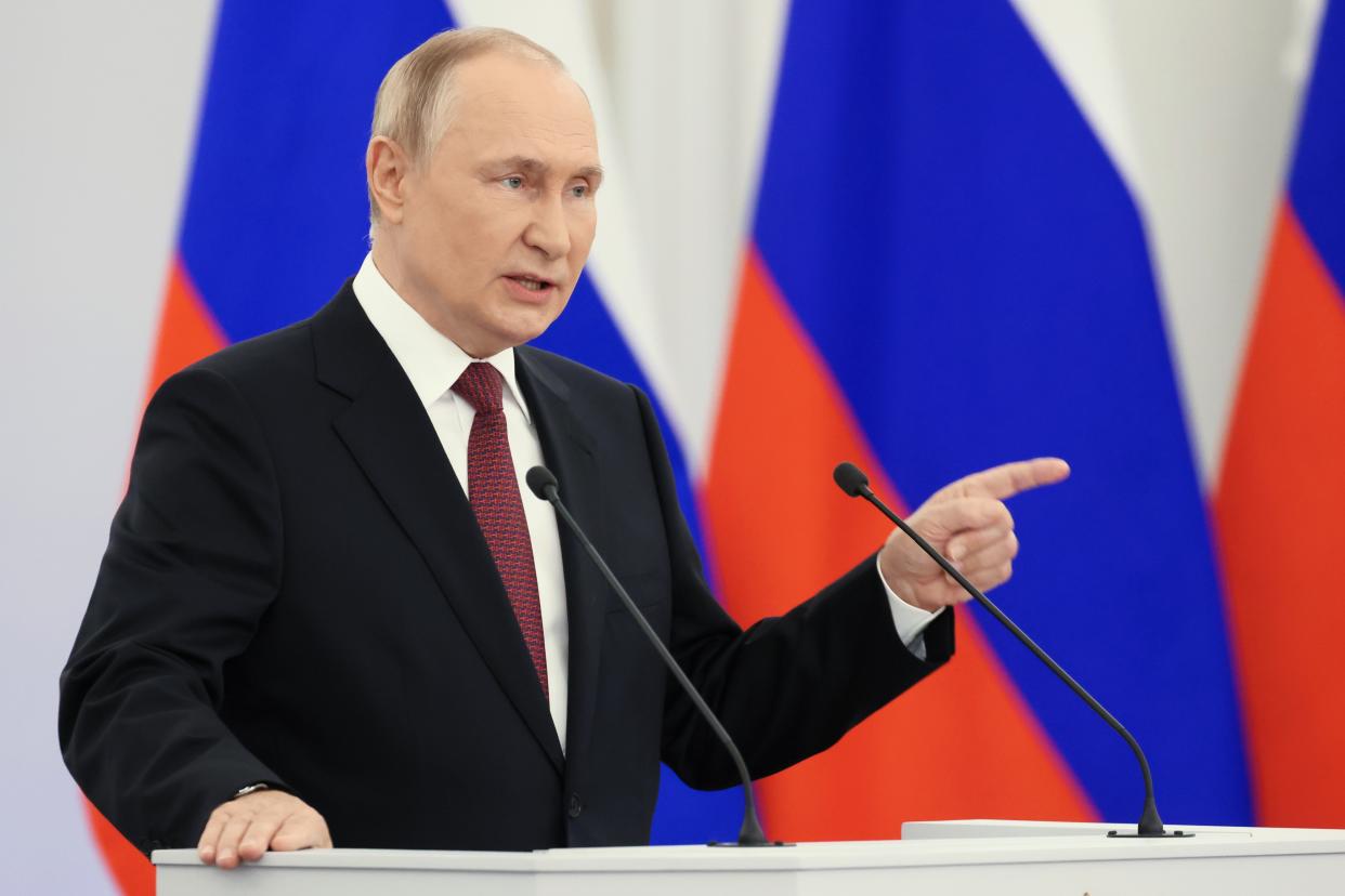 President Vladimir Putin at the microphone, with three Russian flags behind him.