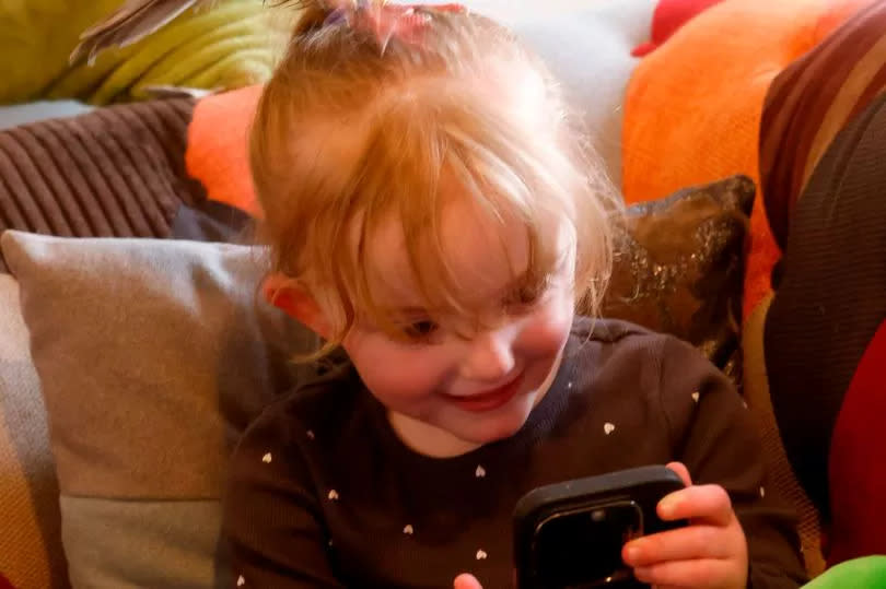 Little Lainey laughs while lying on the sofa playing on her mum's phone while Flori is on Lainey's head