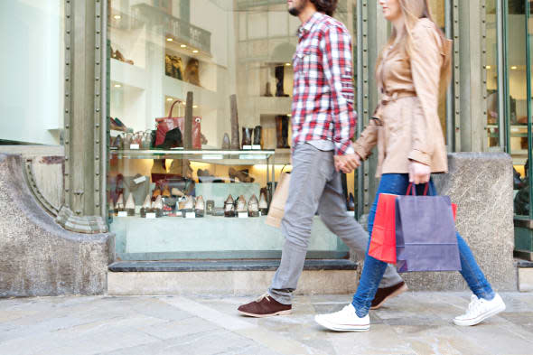 Lower body section of a young tourist couple walking by store windows and holding paper shopping bags in a destination city.