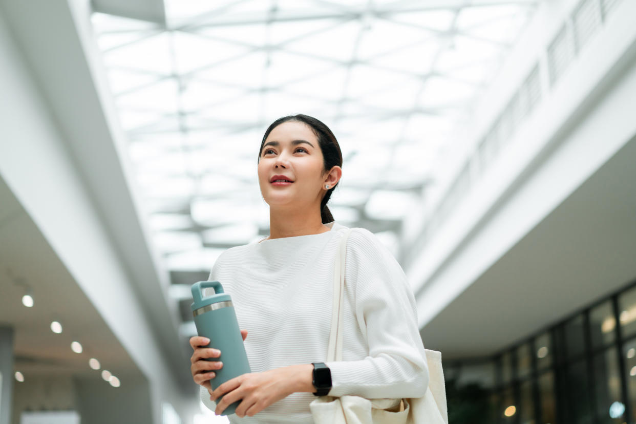 A young woman holding a water bottle and carrying a tote bag walks in an indoor shopping mall.