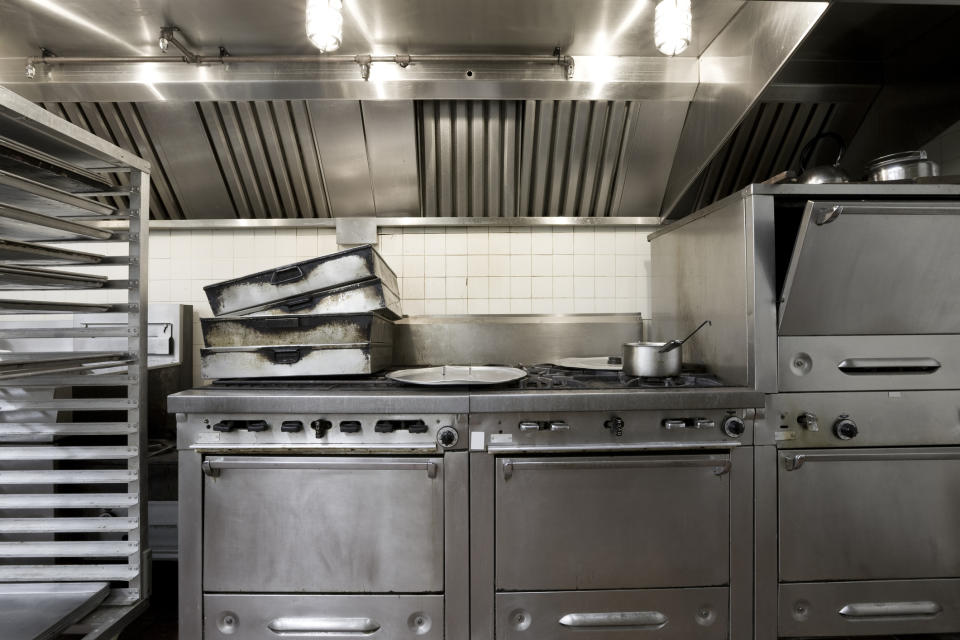 A stainless steel commercial kitchen featuring multiple ovens, a gas stove, stacked empty baking trays, and a saucepan, all set up for professional cooking