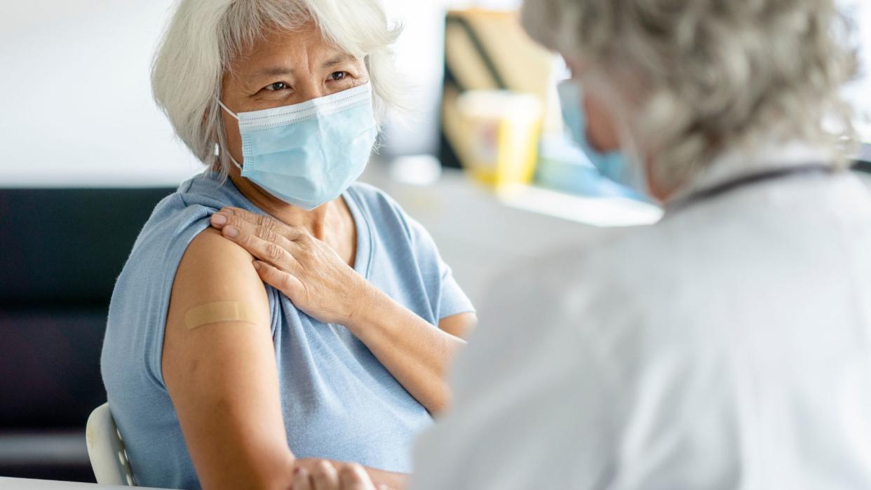  an older woman with white hair sits in a doctor's office with her sleeve rolled up to show a bandage on her arm, as if she'd received a vaccine. She and a doctor, seen in the foreground of the image, are both wearing surgical masks 