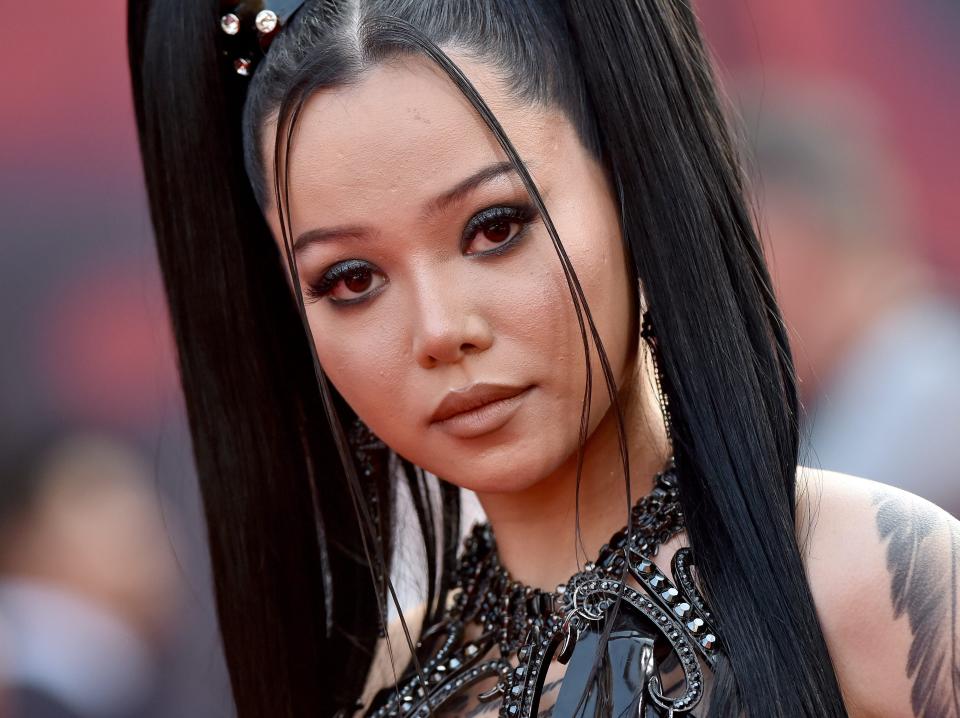 TikTok star bella poarch with her hair in high pigtails, looking at the camera seriously. there's a feather tattoo visible on her shoulder and she's wearing a sleeveless black top with a detailed collar.