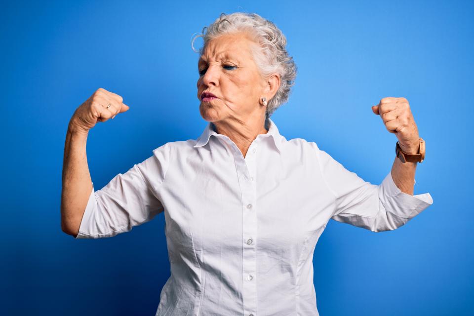 Senior beautiful woman wearing elegant shirt standing over isolated blue background showing arms muscles smiling proud. Fitness concept.