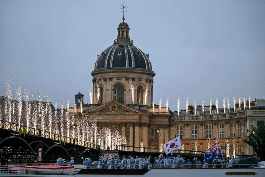 TOPSHOT - Athletes from Cook Islands' delegation (R) and South Korea's delegation sail in a boat along the river Seine past the Pont des Arts footbridge and the Institut de France illuminated with fireworks during the opening ceremony of the Paris 2024 Olympic Games in Paris on July 26, 2024. (Photo by Kirill KUDRYAVTSEV / AFP) (Photo by KIRILL KUDRYAVTSEV/AFP via Getty Images)