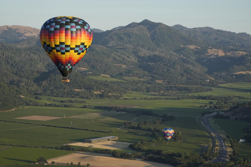 A pair of balloons make their way above the Dominus Estate winery and Highway 29, seen from a Napa Valley Aloft balloon, in Yountville, Calif., Monday, June 19, 2023. This year, wine grapes are thriving after a winter of record amounts of rain fell in California, but a recent trip high above the valley in a hot air balloon revealed miles of lush, green vineyards — the only blemish coming from shadows cast by the balloons themselves. (AP Photo/Eric Risberg)
