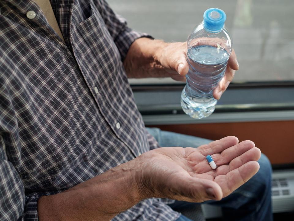 Close-up of unrecognizable aged man in casual shirt holding bottle of water and taking dramamine pill in bus