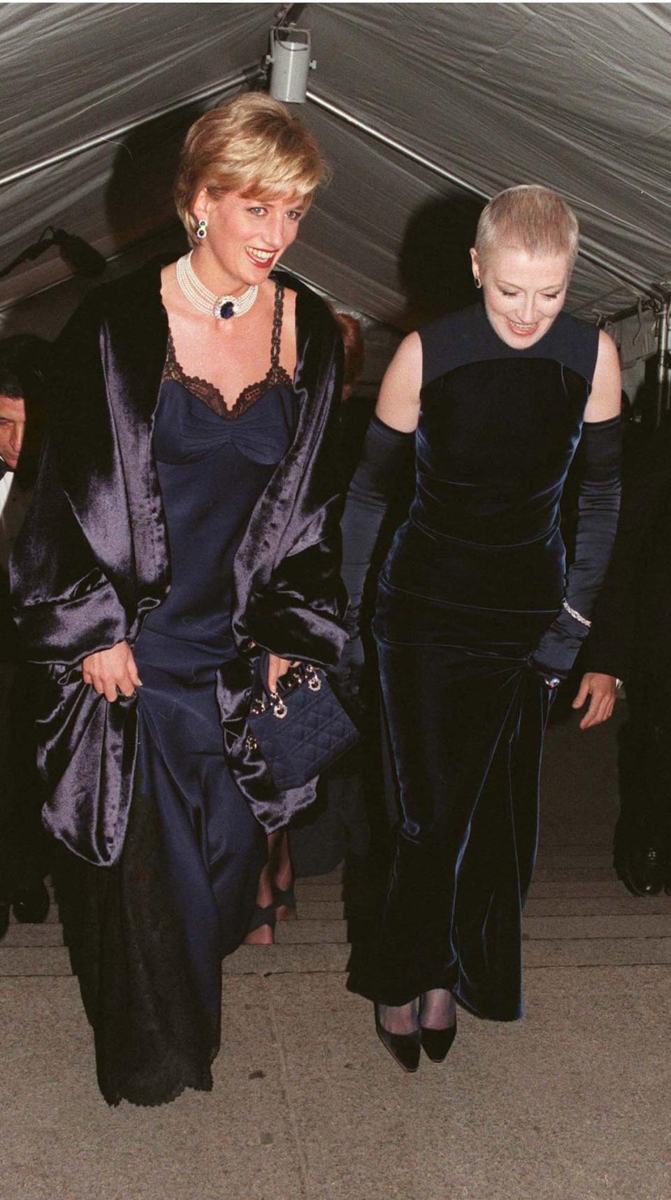 Princess Diana walks up the steps at the 1996 Met Gala, wearing a navy blue slip dress with lace trim and a black shawl. Her friend Liz Tilberis stands next to her, wearing a navy blue velvet gown.