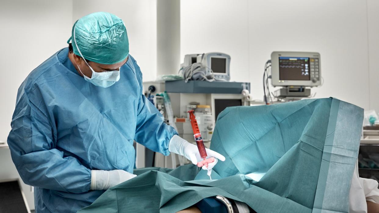  Surgeon monitors the extraction of bone marrow from a patient using a giant syringe in the operating theater. 