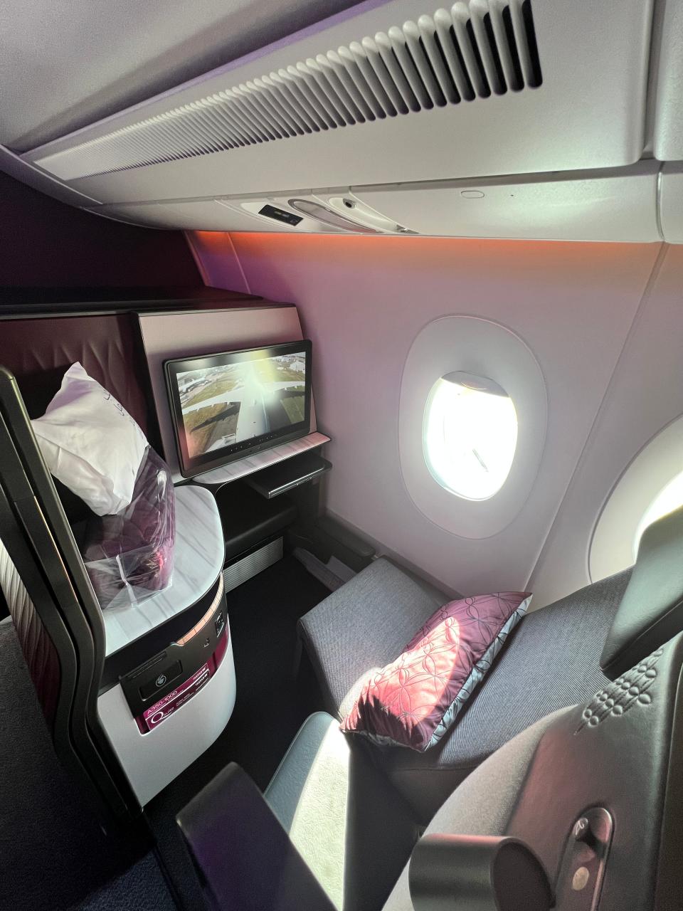 One business class seat on a Qatar Airbus A350, with a burgundy cushion, a window view, and a live feed of the plane on the display screen.