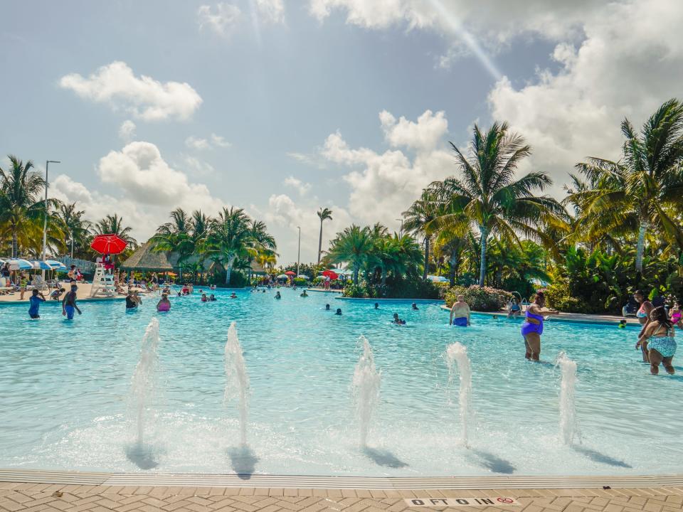 A pool with five small fountains at the entrance at CocoCay with palm trees and cloudy blue skies in the background
