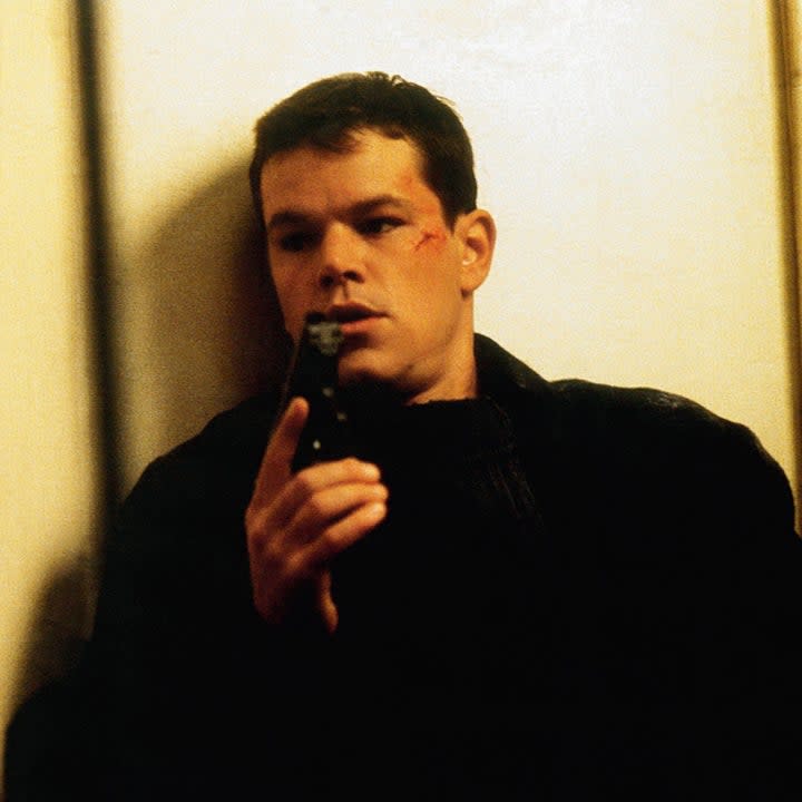 Matt holding a gun as he leans against a wall in a scene from the Bourne films