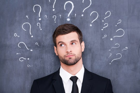 Man standing in front of chalkboard on which are drawn questions marks.