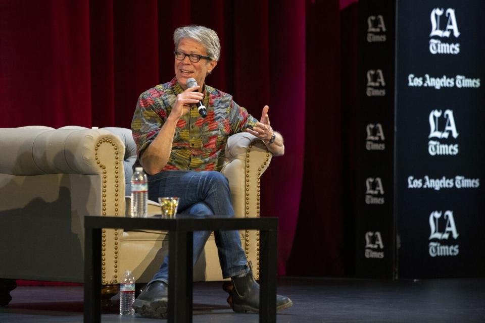 An author with gray hair wearing a flowery shirt, gesturing with a microphone while sitting on a couch.