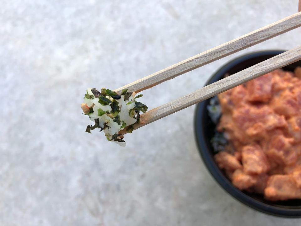 Chopsticks holding some rice covered in furikake, a green seaweed-based topping, with a spicy ahi poke bowl below