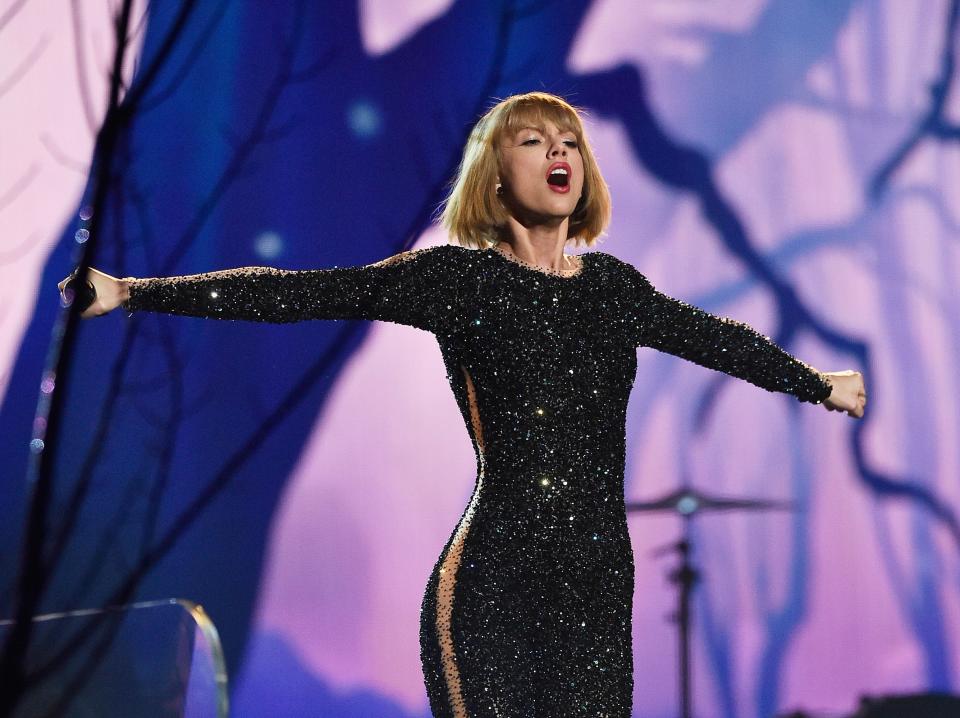 Taylor Swift singing onstage while wearing a figure-hugging black dress and holding her arms out to the side.