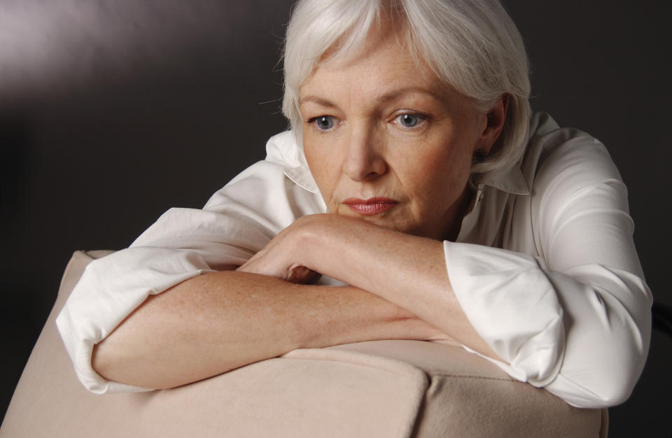 A clearly worried senior woman resting her crossed arms on the back of a chair, and her head on her forearms.