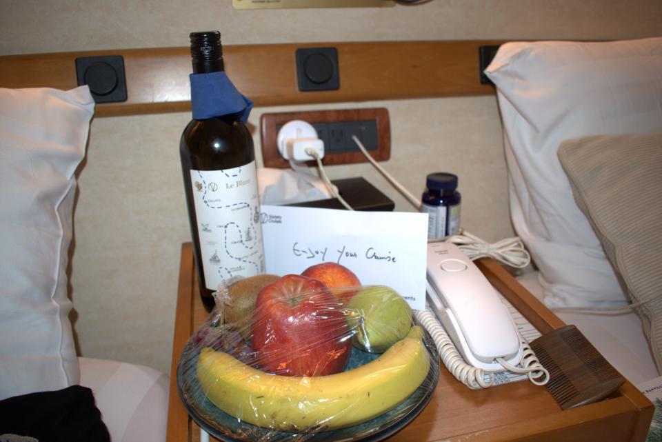 a bottle of wine and bowl of banana, apple, and other fruits next to corded telephone on nightstand