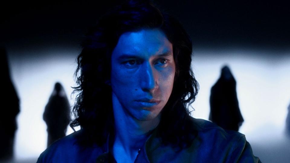 Adam Driver is bathed in blue light and has long dark hair in a scene from Annette.