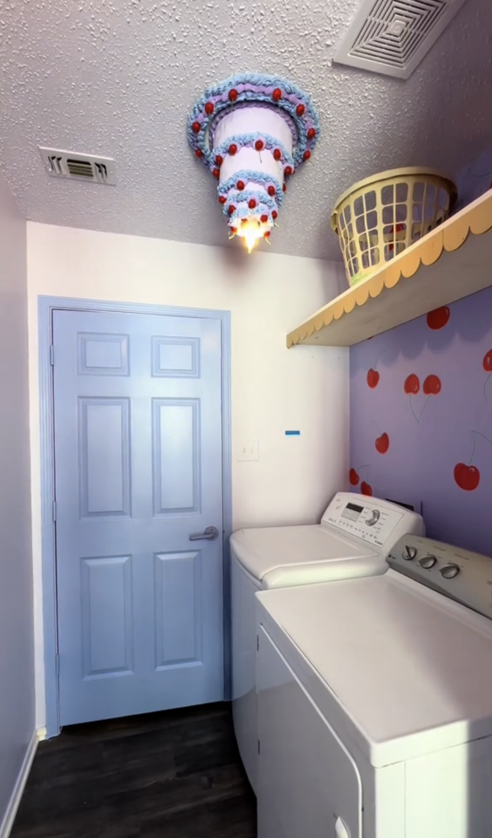 A whimsical laundry room with a unique cake-themed light fixture, a washer, dryer, and walls with hand-painted cherries