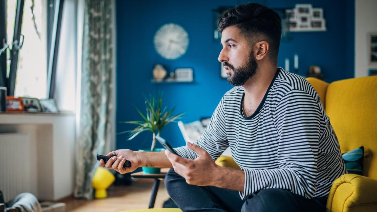 Man sitting home in his armchair, using phone and changing channels.